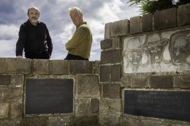 Two men standing behind a stone memorial wall at Penrhos carved with three faces and Welsh inscriptions
