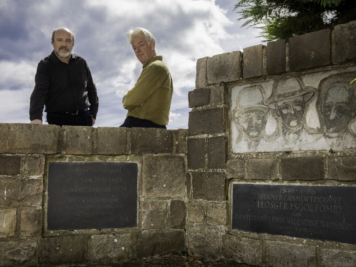 Two men standing behind a stone memorial wall at Penrhos carved with three faces and Welsh inscriptions