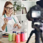 A dressmaker working at a sewing machine in her studio with a camera on a tripod beside her