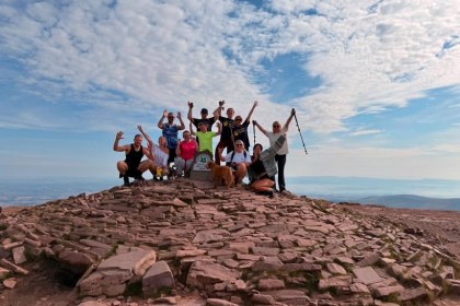 A group of eleven family members and friends waving at the summit cairn of Pen y Fan with a dog, under a blue sky