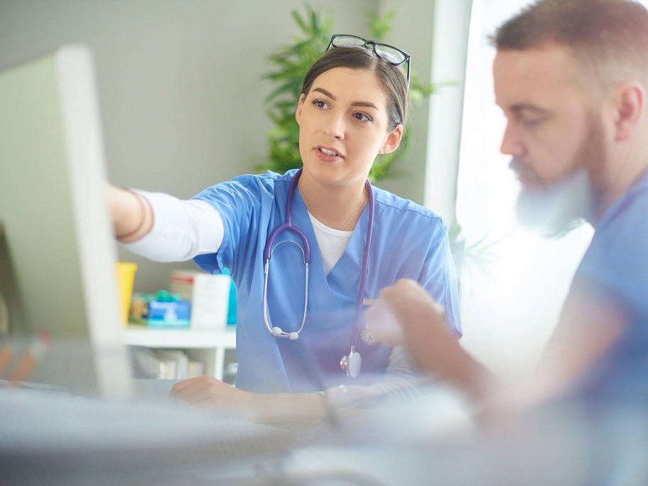 Two clinicians in blue scrubs at a desk, one pointing at a computer screen while the other looks at paperwork