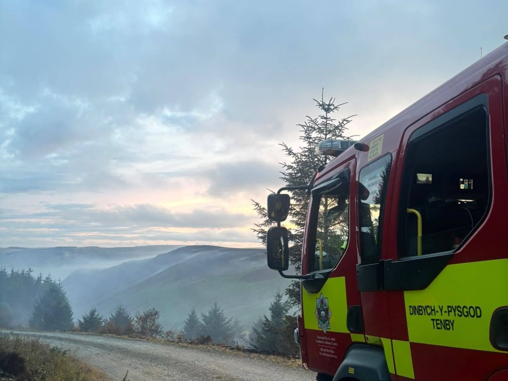 A Mid and West Wales fire appliance from Tenby parked on a forest track at dawn, with mist lying in the valley below