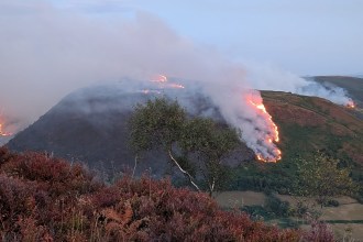 A wildfire burning in lines of flame across a hillside above Rhandirmwyn at dusk, with thick smoke drifting over the valley and farmland below