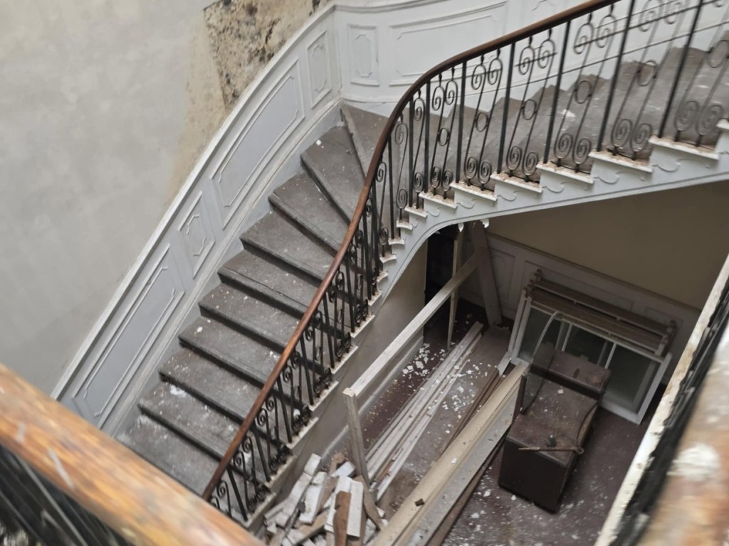 Looking down a curving stone staircase with a slender iron balustrade inside Rheola House, plaster flaking from the walls