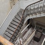 Looking down a curving stone staircase with a slender iron balustrade inside Rheola House, plaster flaking from the walls