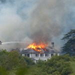 Firefighters on an aerial platform spraying water onto the burning roof of Rheola House, with flames and smoke rising from the Regency manor