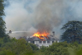 Firefighters on an aerial platform spraying water onto the burning roof of Rheola House, with flames and smoke rising from the Regency manor