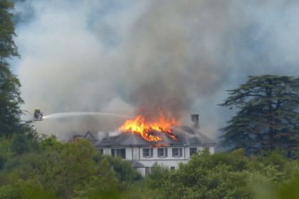 Firefighters on an aerial platform spraying water onto the burning roof of Rheola House, with flames and smoke rising from the Regency manor