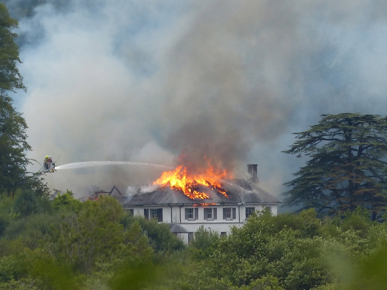 Firefighters on an aerial platform spraying water onto the burning roof of Rheola House, with flames and smoke rising from the Regency manor
