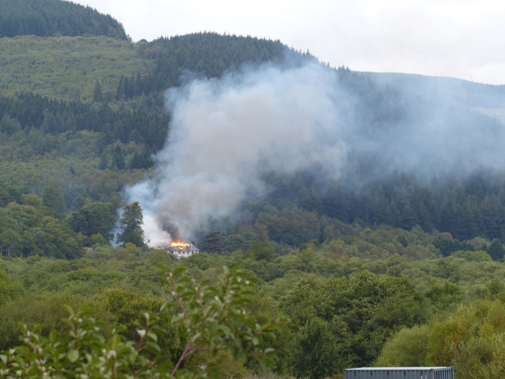A wide view of the wooded Neath Valley with a large plume of smoke rising from Rheola House among the trees
