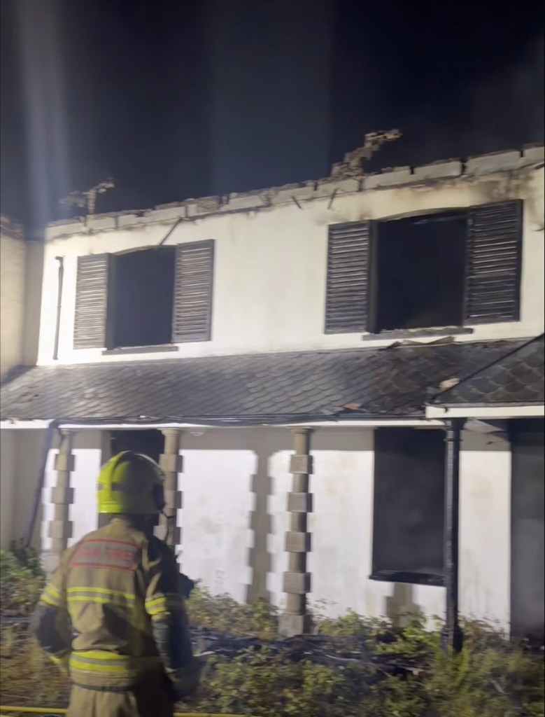 A firefighter standing in front of the fire-damaged white facade of Rheola House at night, smoke drifting from the upper windows