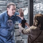 John Partridge as Hannibal Lecter in a blue prison uniform pressing his hand against the glass of his cell, facing Mollie Gallagher as Clarice Starling holding out a black folder