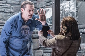 John Partridge as Hannibal Lecter in a blue prison uniform pressing his hand against the glass of his cell, facing Mollie Gallagher as Clarice Starling holding out a black folder