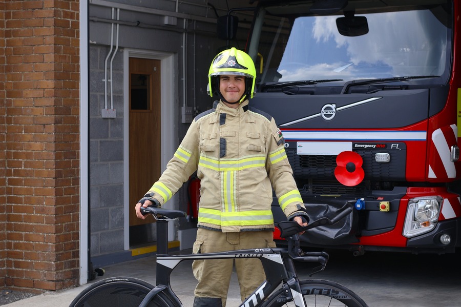 Siôn Jones wearing a firefighter's helmet and kit holding his time trial bike in front of a fire engine with a poppy on its grille