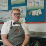Lloyd Henry seated in a school food technology classroom, wearing a grey apron and bow tie
