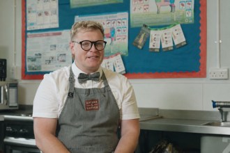 Lloyd Henry seated in a school food technology classroom, wearing a grey apron and bow tie