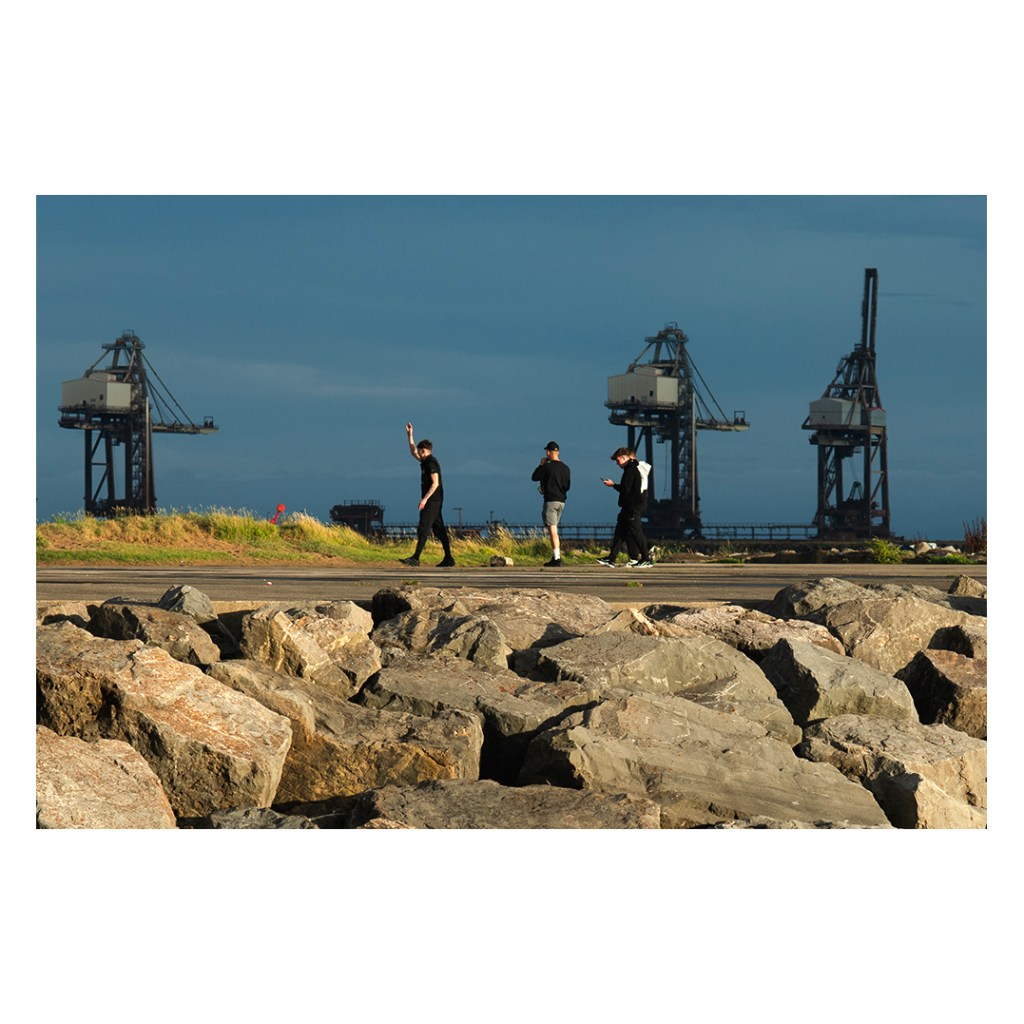 Three young men walking along a coast road with large harbour cranes behind them and boulders in the foreground