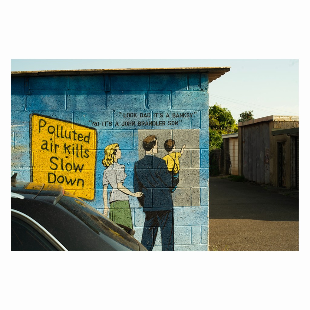 A mural on a blue garage wall reading Polluted air kills, Slow Down, with a painted family looking up at it