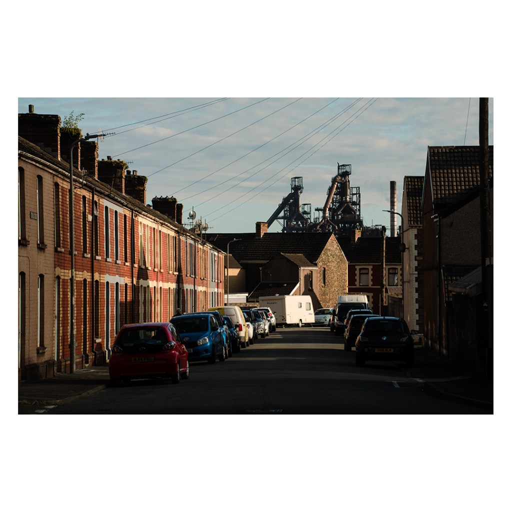 A street of terraced houses in Port Talbot with the blast furnaces visible above the roofline at the end