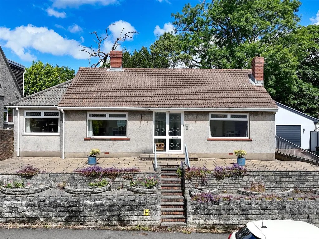 A detached pebbledashed bungalow behind a low stone wall with planted borders and steps up to the door