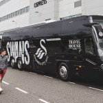 A black Swansea City coach parked outside the Swansea.com Stadium with supporters walking past