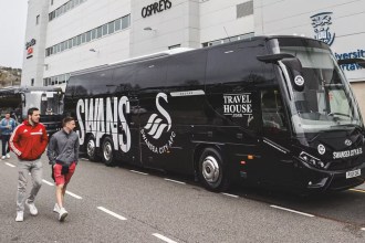 A black Swansea City coach parked outside the Swansea.com Stadium with supporters walking past