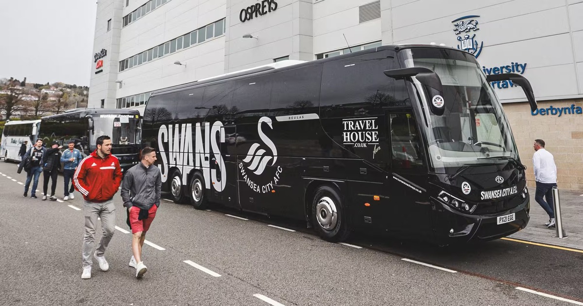 A black Swansea City coach parked outside the Swansea.com Stadium with supporters walking past