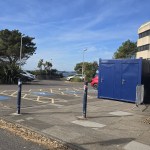 A row of blue portable toilet cabins beside the Civic Centre in a car park with disabled bays, palm trees and Swansea Bay in the background