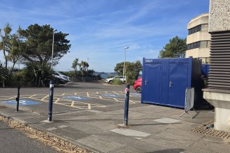 A row of blue portable toilet cabins beside the Civic Centre in a car park with disabled bays, palm trees and Swansea Bay in the background