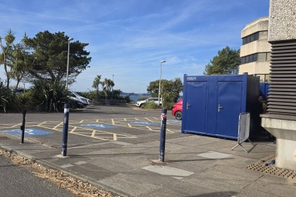 A row of blue portable toilet cabins beside the Civic Centre in a car park with disabled bays, palm trees and Swansea Bay in the background