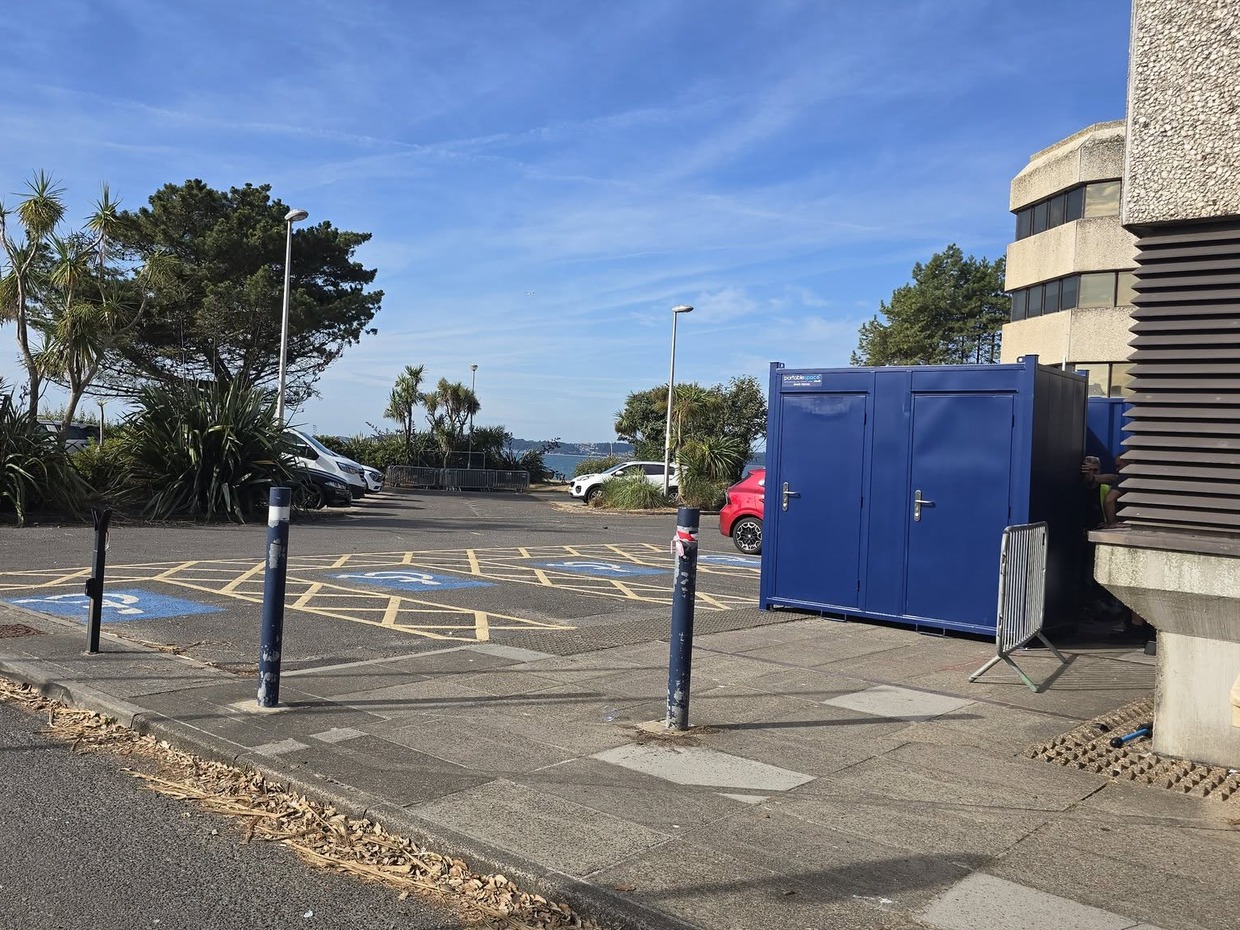 A row of blue portable toilet cabins beside the Civic Centre in a car park with disabled bays, palm trees and Swansea Bay in the background