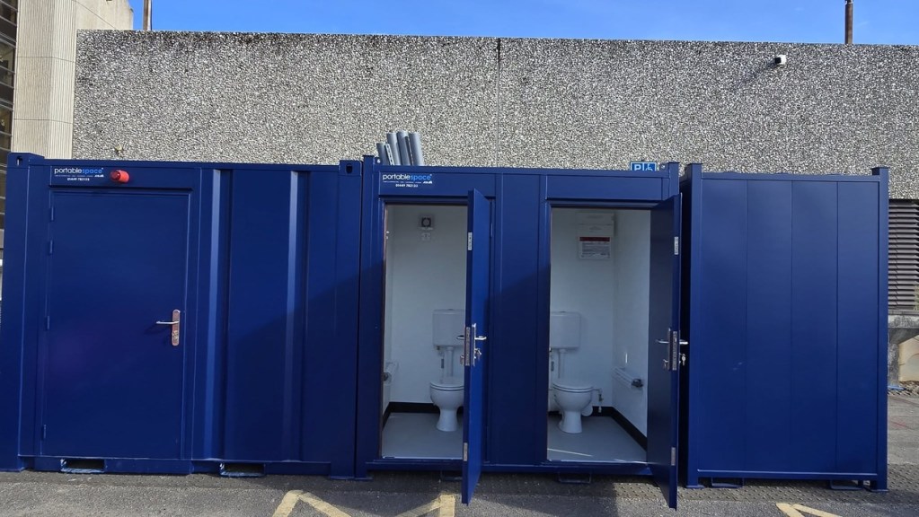 Blue portable toilet cabins with two doors open showing white toilets inside, against the pebble-dashed wall of the Civic Centre
