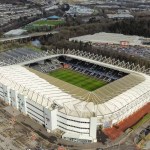 Aerial view of the Swansea.com Stadium in Landore, Swansea, with its white steel roof frame, the pitch visible below, and the River Tawe and retail park beyond