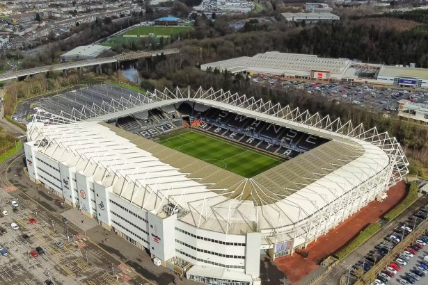 Aerial view of the Swansea.com Stadium in Landore, Swansea, with its white steel roof frame, the pitch visible below, and the River Tawe and retail park beyond