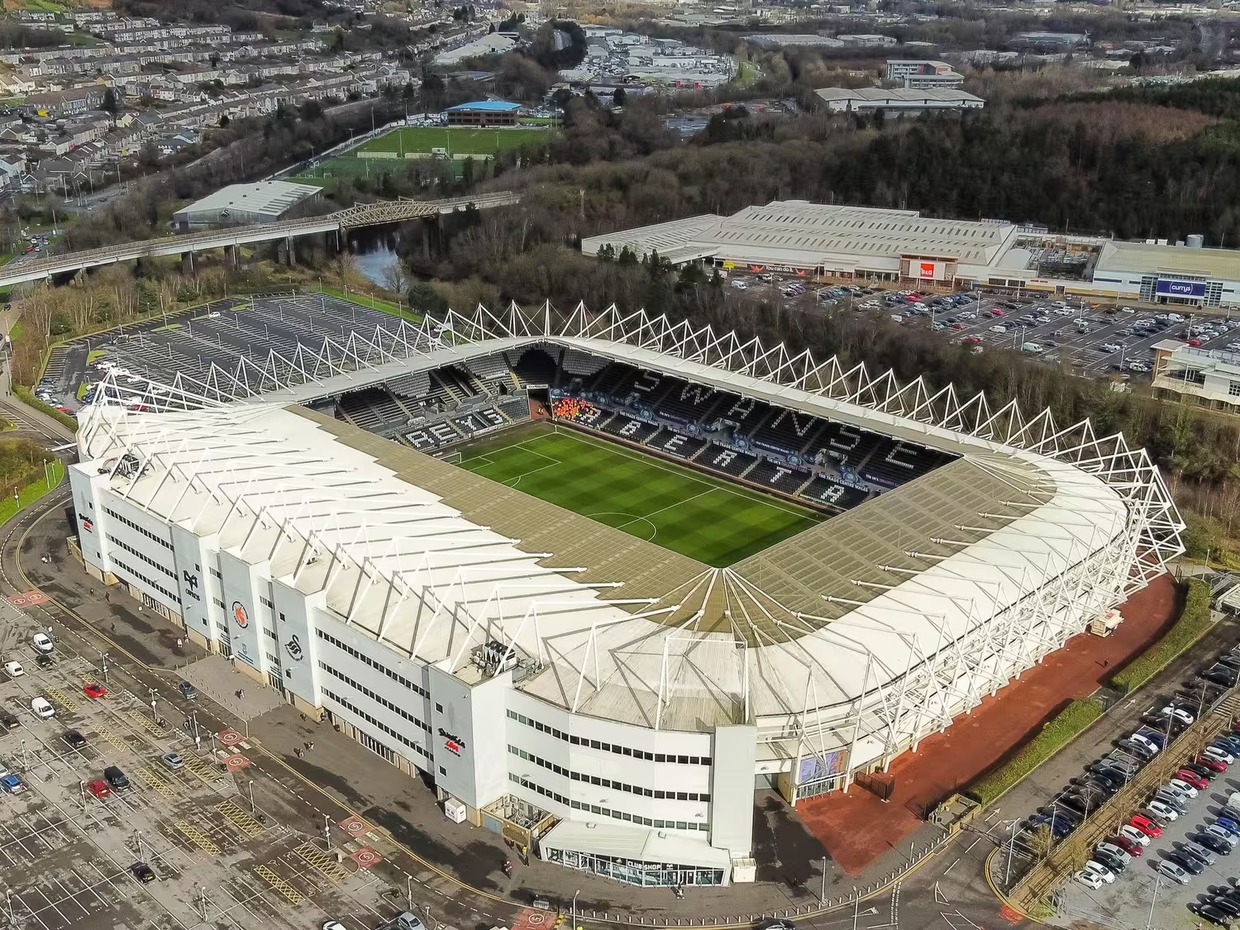 Aerial view of the Swansea.com Stadium in Landore, Swansea, with its white steel roof frame, the pitch visible below, and the River Tawe and retail park beyond