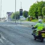 A police motorcyclist rides past a digital rider feedback sign on Carmarthen Road, Swansea, displaying 29 in green with a motorbike symbol