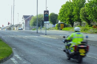 A police motorcyclist rides past a digital rider feedback sign on Carmarthen Road, Swansea, displaying 29 in green with a motorbike symbol