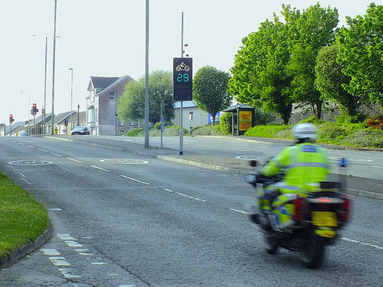A police motorcyclist rides past a digital rider feedback sign on Carmarthen Road, Swansea, displaying 29 in green with a motorbike symbol