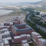 Aerial view of the red-brick Bay Campus buildings between the beach at Crymlyn Burrows and Fabian Way, with Kilvey Hill beyond