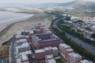 Aerial view of the red-brick Bay Campus buildings between the beach at Crymlyn Burrows and Fabian Way, with Kilvey Hill beyond