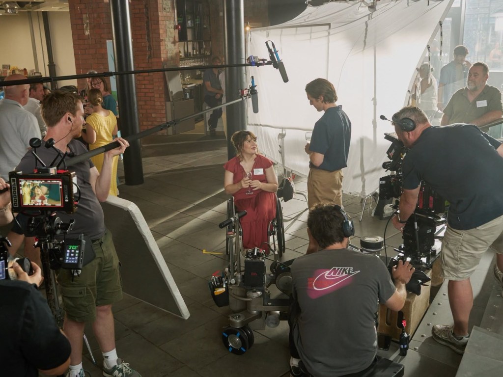 Film crew with cameras and boom microphones filming Ruth Madeley in a wheelchair during an indoor scene for The Rapture