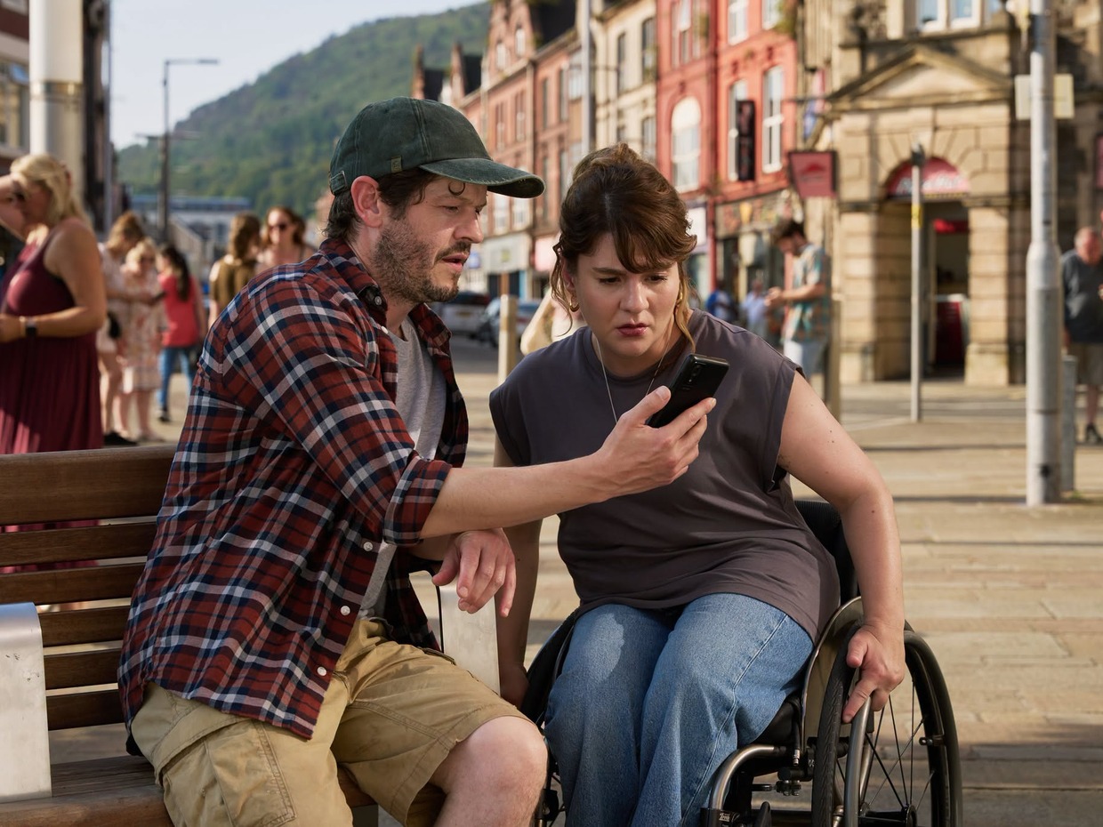 Iwan Rheon and Ruth Madeley looking at a phone on a bench in Port Talbot town centre during filming of The Rapture