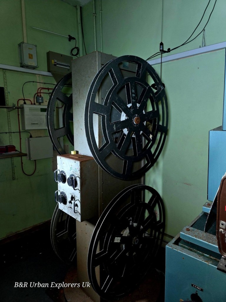 A film projector with large reels in the green-walled projection room of the derelict cinema