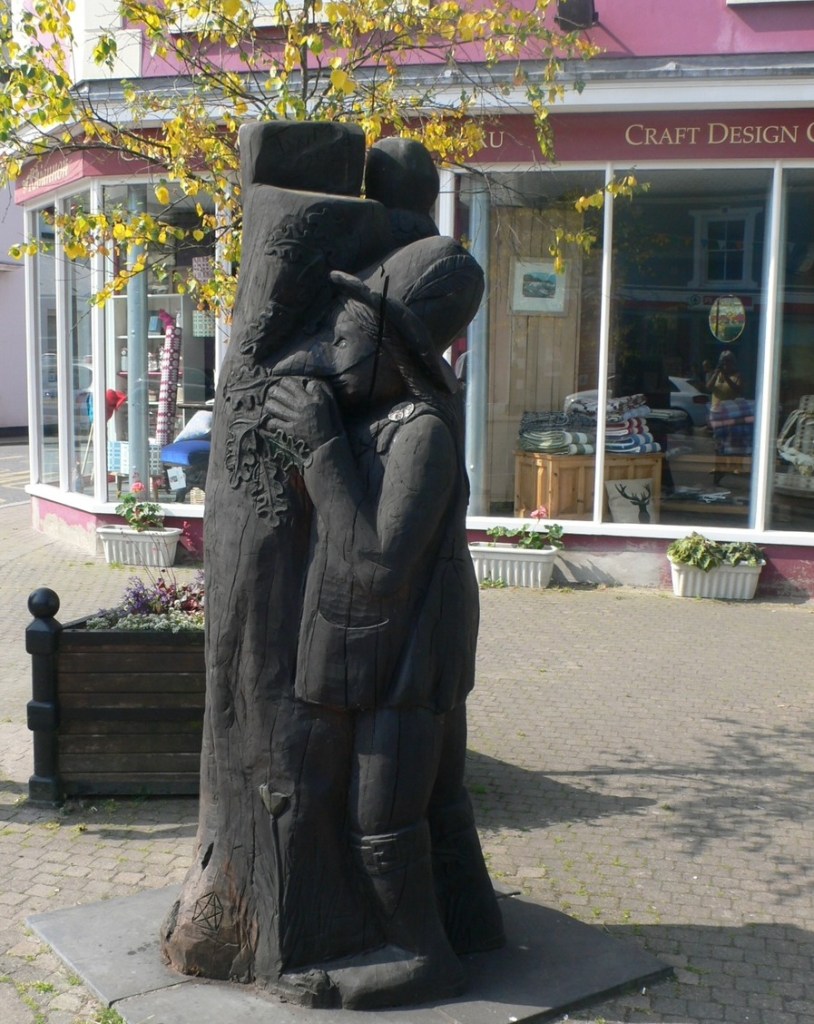 A dark carved wooden statue of Twm Siôn Cati beside a tree trunk on Tregaron Square, with a craft shop behind