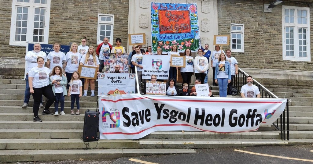 Parents, children and campaigners on the steps of County Hall behind a large Save Ysgol Heol Goffa banner, holding placards and wearing t-shirts printed with photographs of pupils