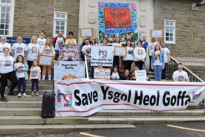 Parents, children and campaigners on the steps of County Hall behind a large Save Ysgol Heol Goffa banner, holding placards and wearing t-shirts printed with photographs of pupils