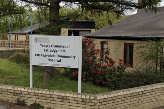 The entrance sign and low brick buildings of Ystradgynlais Community Hospital, with the bilingual Ysbyty Cymunedol Ystradgynlais sign in front