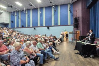 A packed hall of seated residents viewed from the back as David Chadwick MP speaks from the stage