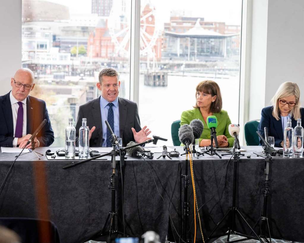 Four party representatives sit behind microphones at a press conference overlooking Cardiff Bay.