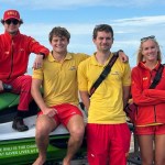 Six RNLI lifeguards in red and yellow uniforms posing with a rescue watercraft on the promenade at Aberavon Beach